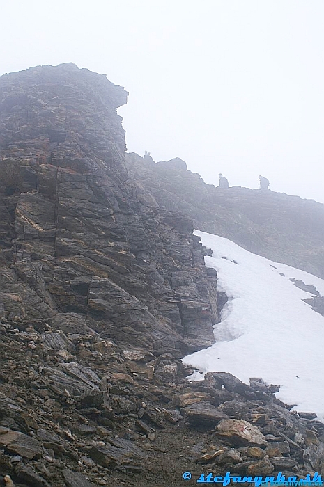 Spielmann. Za skalou v ľavej časti sa nachádza vrcholový kríž. Lano začína pod ľavým človekom na úrovni horného okraja snehu