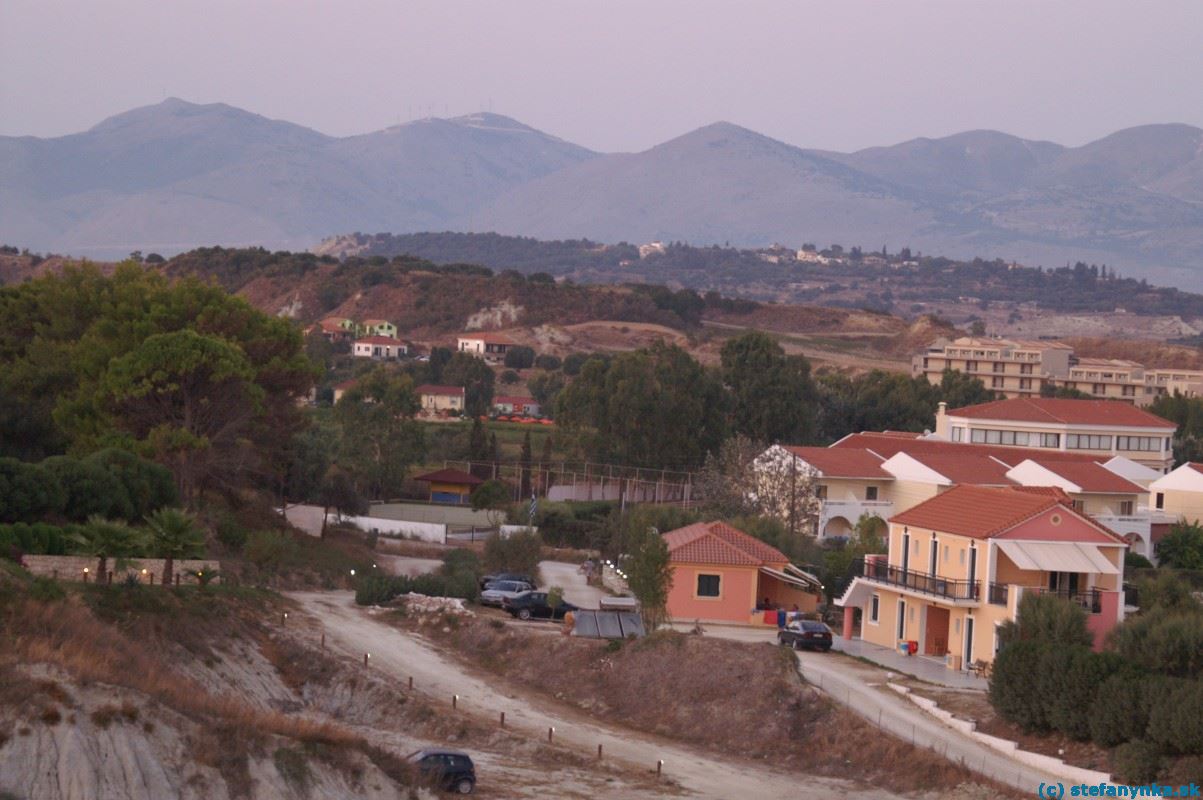 Lixouri-Xi, Kefalónia. Stredisko Xi z útesov. Vpravo trčí recepcia s jedálňou hotela Kefalonia palace. Za recepciou je vidieť budovy nového hotela (Apolonia alebo tak nejako) s nevýraznou farbou strechy. Približne v strede obrázka je budova autopôžičovne (červená strecha a rad červených áut pred budovou)