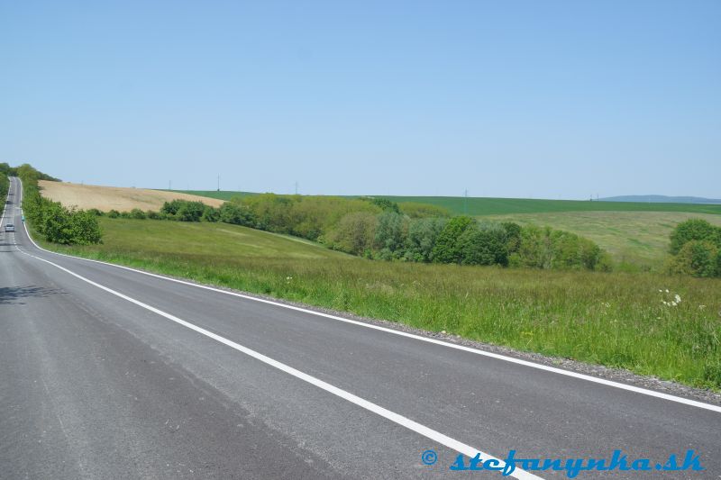 Cesta od Miléniového kríža (za chrbtom) na Radošovce. Za autom je odbočka doprava k Prameňu (medza a rozhranie poľa a lúky s kríkmi). Na vrchu asfaltky je odbočka doľava do kúpeľov Smrdáky